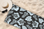 Black towel with white floral pattern laid on sand, accompanied by a sun hat and sunglasses; stylish beach essentials.