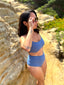 Side view of person wearing cornflower blue swim top and mid-rise bottom, standing against sandy backdrop; elegant swimwear detail.