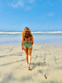 Woman walking toward the ocean wearing a green bikini, criss-cross back tie and mid-rise fit bottom.