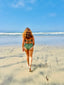 Woman walking toward the ocean wearing a green bikini, criss-cross back tie and mid-rise fit bottom.
