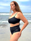 Side view of black swimsuit with hidden-support top and mid-rise bottom, worn on sandy beach; modern, supportive swimwear style.