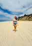 Woman walking on sandy beach wearing a blue bikini, showcasing Vie Swimwear.