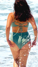 Green swimsuit with crisscross tie-back top and mid-rise bottom, worn by a person standing in ocean water; vibrant swimwear style.