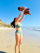 Person wearing green swimsuit with hidden-support top and mid-rise bottom, standing on a sandy beach while holding a baby.