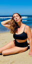 Black swimsuit with hidden-support top and mid-rise bottom, worn while sitting on a sandy beach; relaxed swimwear style.