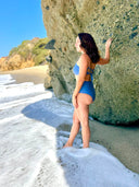 Side view of person in blue swimsuit with criss-cross back string top and mid-rise bottom, standing by rock wall on the beach.