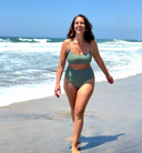 Person wearing green swimsuit with mid-rise top and bottom, walking along a sandy beach; casual swimwear style.