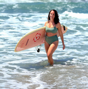 Person wearing green swimsuit with mid-rise top and bottom, standing in ocean water holding a surfboard; active swimwear vibe.