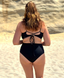 Black mid-rise swimsuit bottom with top back tie detail, worn on a sandy beach; minimalist swimwear style.