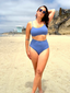 Woman wearing cornflower blue bikini top and high-waisted bottoms, standing on a sandy beach; fresh beachwear style.