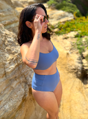 Side view of person wearing blue swimsuit with hidden-support top and mid-rise bottom, standing on a rock wall; sleek swimwear style.
