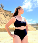 Black swimsuit set with hidden-support top and mid-rise bottom, worn on a sandy beach; clean, modern swimwear design.