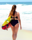 Black mid-rise swimsuit bottom with back tie, worn on a sandy beach beside a surfboard; sporty, stylish swimwear moment.