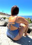 Person in blue swimsuit sitting on beach rocks, wearing criss-cross back string top and mid-rise bottom bikini; relaxed seaside style.
