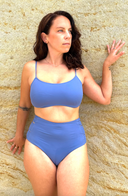 Person wearing cornflower blue swim top and mid-rise bottom, standing against a sandy backdrop; fresh, vibrant swimwear look.