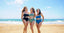 Three women smiling on a sandy beach in stylish, supportive Vie Swimwear — black, blue, and green suits designed for confidence and comfort.